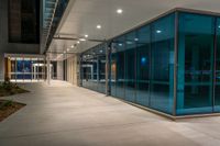 a sidewalk outside a large glass building next to another one with lights at night on them