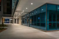 a sidewalk outside a large glass building next to another one with lights at night on them