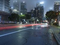 a street at night with fog covered city buildings in the background and car lights on