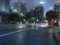 a street at night with fog covered city buildings in the background and car lights on