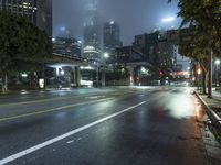a street at night with fog covered city buildings in the background and car lights on