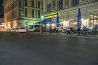 a street is lit with outdoor tables and umbrellas and cars are parked in front of the restaurant