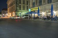 a street is lit with outdoor tables and umbrellas and cars are parked in front of the restaurant