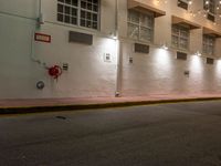 an empty city street at night with lights on the buildings behind it and two fire hydrants along the curb