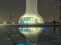 a water fountain at night with lights reflecting off it's surface and in front of a tall modern building