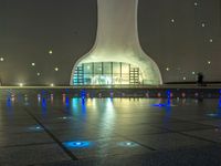 a water fountain at night with lights reflecting off it's surface and in front of a tall modern building