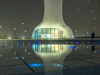 a water fountain at night with lights reflecting off it's surface and in front of a tall modern building