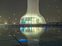 a water fountain at night with lights reflecting off it's surface and in front of a tall modern building