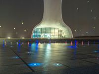 a water fountain at night with lights reflecting off it's surface and in front of a tall modern building