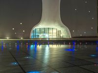 a water fountain at night with lights reflecting off it's surface and in front of a tall modern building