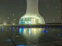 a water fountain at night with lights reflecting off it's surface and in front of a tall modern building