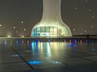 a water fountain at night with lights reflecting off it's surface and in front of a tall modern building