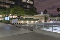 an empty sidewalk in front of a tall building at night with people walking around it