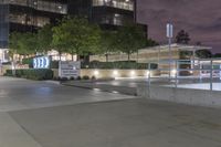 an empty sidewalk in front of a tall building at night with people walking around it