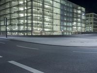 an empty road in front of a glass building at night with lights on it's windows