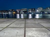 the night lights and grass line the concrete sidewalks by the water and buildings, overlooking the city