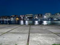 the night lights and grass line the concrete sidewalks by the water and buildings, overlooking the city