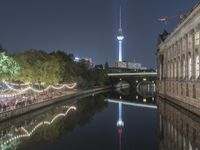 people are sitting at tables near the water by a river and a tall building with a tv tower in the background