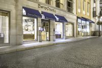 this is the street with several store front windows at night, on which there are multiple different shops along with one empty sidewalk