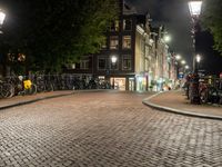 a group of bikes parked on side of a brick road at night with lights on