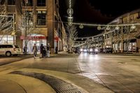 an old street filled with people and parked cars at night time, surrounded by holiday lights