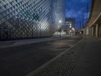 a street with stone sidewalks in front of a glassy building at night on the corner