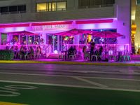 several people sitting at tables outside of a restaurant in pink lights at night time and outside of it is a building with green shutters