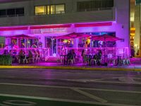 several people sitting at tables outside of a restaurant in pink lights at night time and outside of it is a building with green shutters