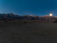 a view of mountains at night with the moon rising above them and a desert area in foreground