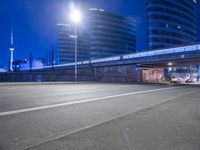 an empty street with buildings in the background at night and a road crossing above it
