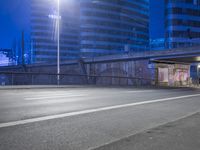 an empty street with buildings in the background at night and a road crossing above it