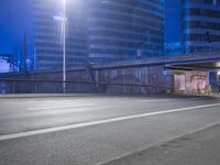 an empty street with buildings in the background at night and a road crossing above it