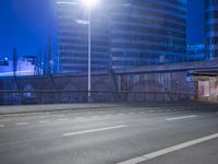 an empty street with buildings in the background at night and a road crossing above it