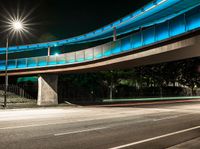 a very long blue bridge over a highway near trees and a street light lit up on the side of it