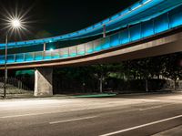 a very long blue bridge over a highway near trees and a street light lit up on the side of it