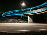 a very long blue bridge over a highway near trees and a street light lit up on the side of it