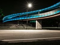 a very long blue bridge over a highway near trees and a street light lit up on the side of it