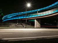 a very long blue bridge over a highway near trees and a street light lit up on the side of it