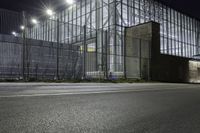 a street near a building with steel fencing around it at night hours, while light shines off windows