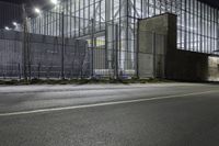 a street near a building with steel fencing around it at night hours, while light shines off windows