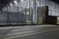 a street near a building with steel fencing around it at night hours, while light shines off windows