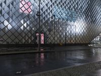 people on the sidewalk walking on a wet surface with a large glass building in the background