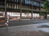 a city sidewalk next to a building filled with lots of windows and wooden benches,