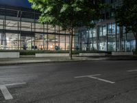 an empty road and some windows in the night time near trees and bushes, with a glass building