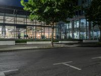 an empty road and some windows in the night time near trees and bushes, with a glass building