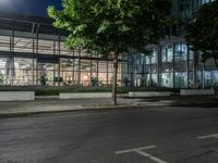 an empty road and some windows in the night time near trees and bushes, with a glass building