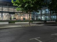 an empty road and some windows in the night time near trees and bushes, with a glass building