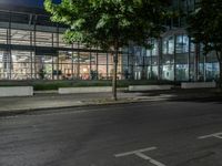 an empty road and some windows in the night time near trees and bushes, with a glass building