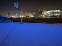 an empty basketball court next to a city at night time, at a distance is a neon lit skyscraper