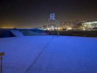 an empty basketball court next to a city at night time, at a distance is a neon lit skyscraper